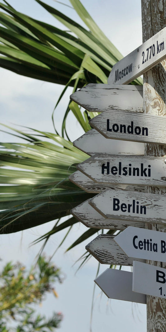 Ein hölzerner Wegweiser vor Palmen zeigt Entfernungen zu verschiedenen Städten: Moskau (2.770 km), London (3.840 km), Helsinki (4.081 km), Berlin (3.027 km), Beirut (1.460 km) und zum Cettia Beach Resort. Die Schilder sind wettergegerbt und zeigen in unterschiedliche Richtungen.

Kurze Version (z. B. für Screenreader mit Zeichenlimit):
Hölzerner Wegweiser vor Palmen zeigt Entfernungen zu Städten wie Moskau, London und Beirut.