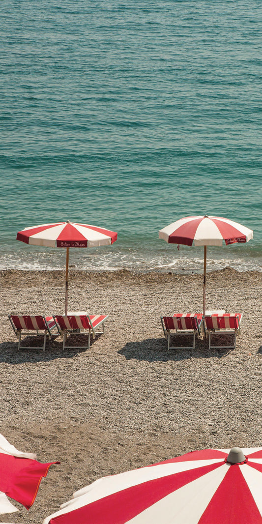 Reihe von rot-weißen Sonnenschirmen und passenden Liegestühlen an einem Kiesstrand mit Blick auf das ruhige, türkisfarbene Meer bei sonnigem Wetter.