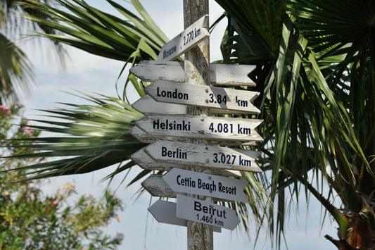 Ein hölzerner Wegweiser vor Palmen zeigt Entfernungen zu verschiedenen Städten: Moskau (2.770 km), London (3.840 km), Helsinki (4.081 km), Berlin (3.027 km), Beirut (1.460 km) und zum Cettia Beach Resort. Die Schilder sind wettergegerbt und zeigen in unterschiedliche Richtungen.

Kurze Version (z. B. für Screenreader mit Zeichenlimit):
Hölzerner Wegweiser vor Palmen zeigt Entfernungen zu Städten wie Moskau, London und Beirut.