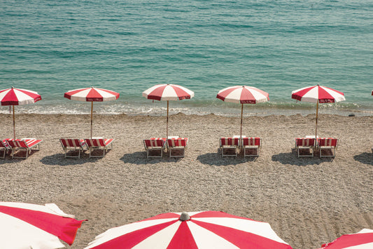 Reihe von rot-weißen Sonnenschirmen und passenden Liegestühlen an einem Kiesstrand mit Blick auf das ruhige, türkisfarbene Meer bei sonnigem Wetter.